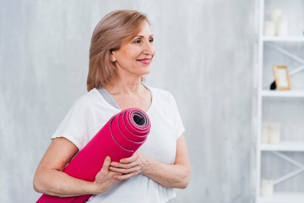 Mujer bonita feliz que se coloca en el rollo casero de la tenencia de la estera de la yoga disponible