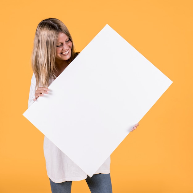 Mujer bonita feliz que sostiene la cartulina blanca en blanco contra el papel pintado amarillo