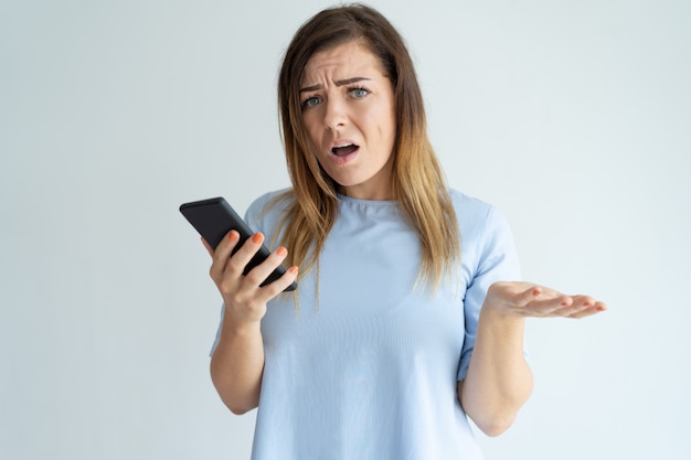 Mujer bonita indignada que lanza la mano, sosteniendo smartphone y mirando la cámara.