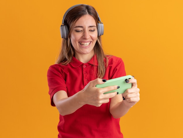 Mujer bonita joven alegre en auriculares sostiene y mira el teléfono aislado en la pared naranja