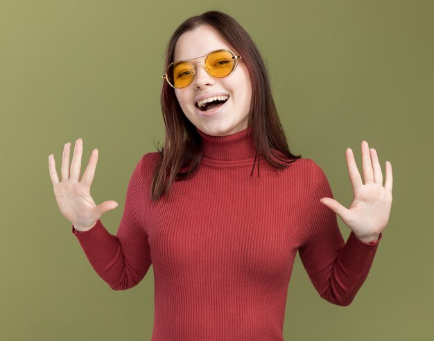 Mujer bonita joven alegre con gafas de sol mirando al frente mostrando las manos vacías aisladas en la pared verde oliva