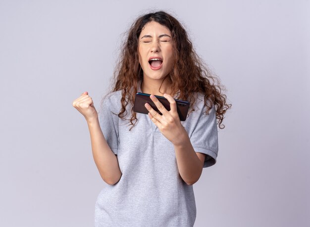 Mujer bonita joven alegre que sostiene el teléfono móvil haciendo sí el gesto con los ojos cerrados aislado en la pared blanca con espacio de copia