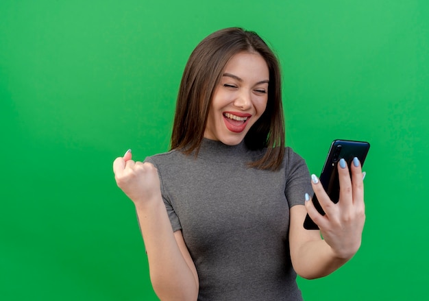 Mujer bonita joven alegre sosteniendo y mirando móvil y haciendo sí gesto teléfono aislado sobre fondo verde con espacio de copia