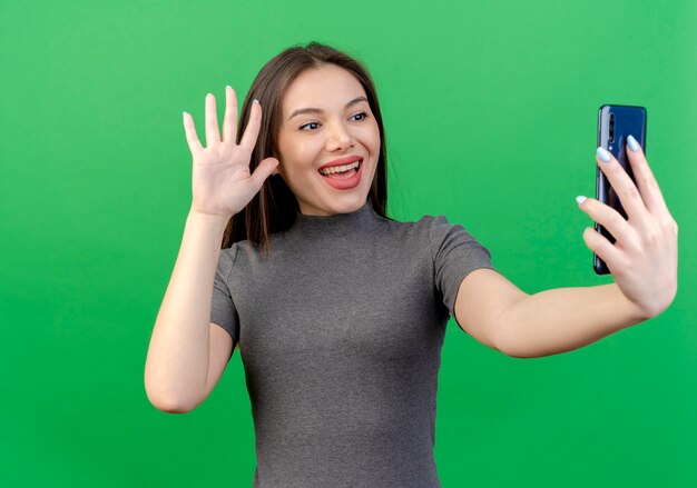 Mujer bonita joven alegre sosteniendo mirando y saludando al teléfono móvil aislado sobre fondo verde