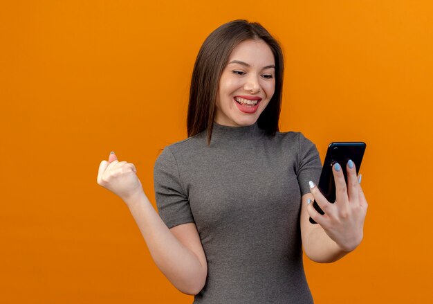 Mujer bonita joven alegre sosteniendo y mirando el teléfono móvil y apretando el puño aislado sobre fondo naranja