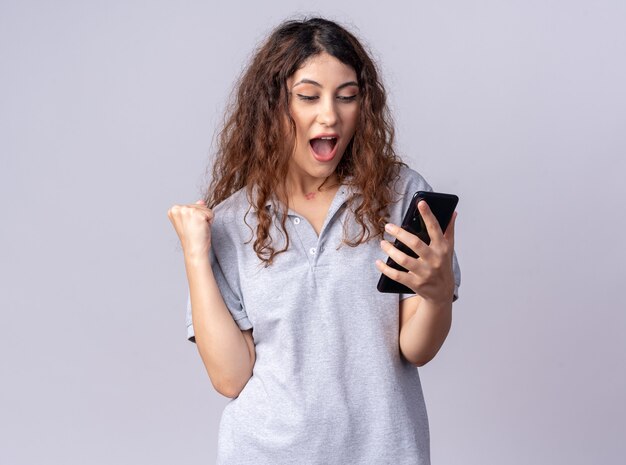 Mujer bonita joven alegre sosteniendo y mirando el teléfono móvil haciendo gesto sí aislado en la pared blanca con espacio de copia