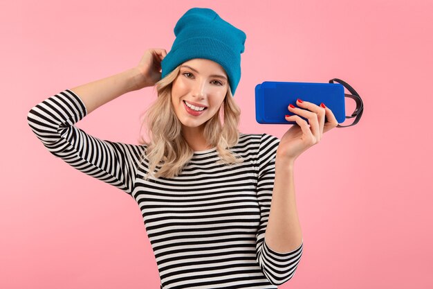 Mujer bonita joven con altavoz inalámbrico escuchando música con camisa a rayas y sombrero azul sonriendo feliz estado de ánimo positivo posando sobre fondo rosa aislado