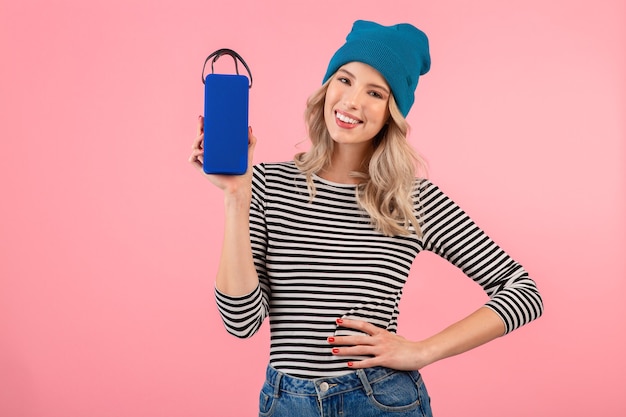 Mujer bonita joven con altavoz inalámbrico escuchando música con camisa a rayas y sombrero azul sonriendo feliz estado de ánimo positivo posando sobre fondo rosa