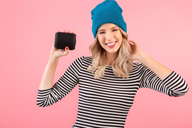 Mujer bonita joven con altavoz inalámbrico escuchando música con camisa a rayas y sombrero azul sonriendo feliz estado de ánimo positivo posando sobre fondo rosa