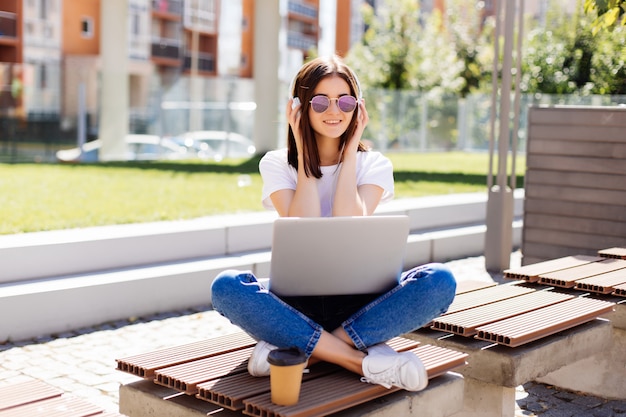 Foto gratuita mujer bonita joven con los auriculares que se sientan en banco en el parque, usando la computadora portátil, establecimiento de una red en línea en inalámbrico.