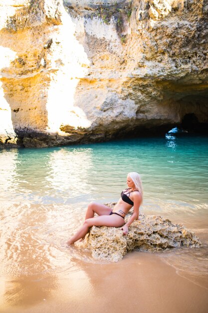 Mujer bonita joven en bikini negro posando sobre una arena de rocas cerca del mar