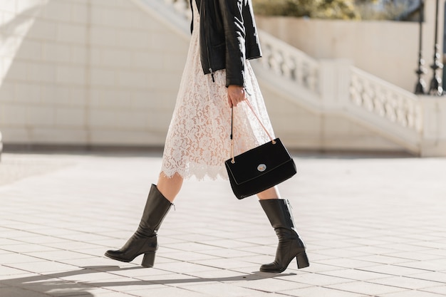 Mujer bonita joven caminando en la calle en traje de moda, sosteniendo el bolso, vestida con chaqueta de cuero negro y vestido de encaje blanco, estilo primavera otoño, clima cálido y soleado, look romántico