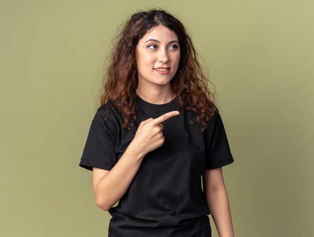 Mujer bonita joven complacida mirando y apuntando al lado aislado en la pared verde oliva con espacio de copia