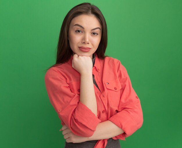 Mujer bonita joven confiada que pone la mano en la barbilla mirando al frente aislado en la pared verde