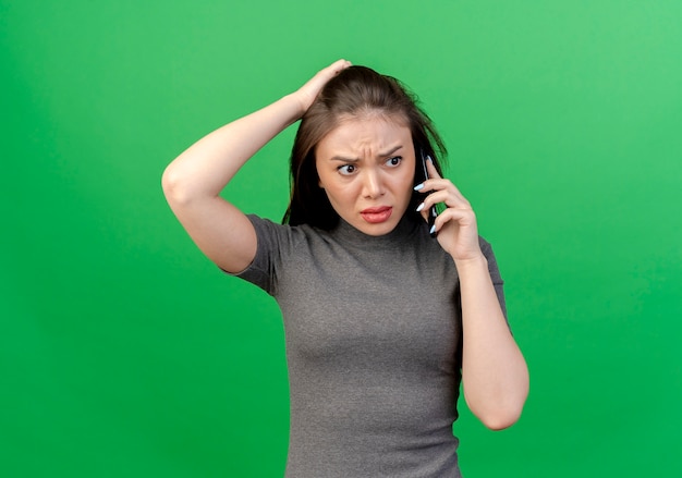 Mujer bonita joven disgustada hablando por teléfono mirando al lado poniendo la mano en la cabeza aislada sobre fondo verde con espacio de copia