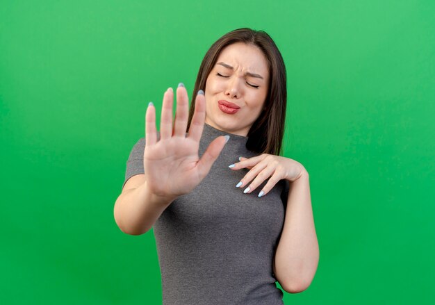 Mujer bonita joven disgustada manteniendo la mano en el pecho y gesticulando parada en la cámara con los ojos cerrados aislado sobre fondo verde con espacio de copia