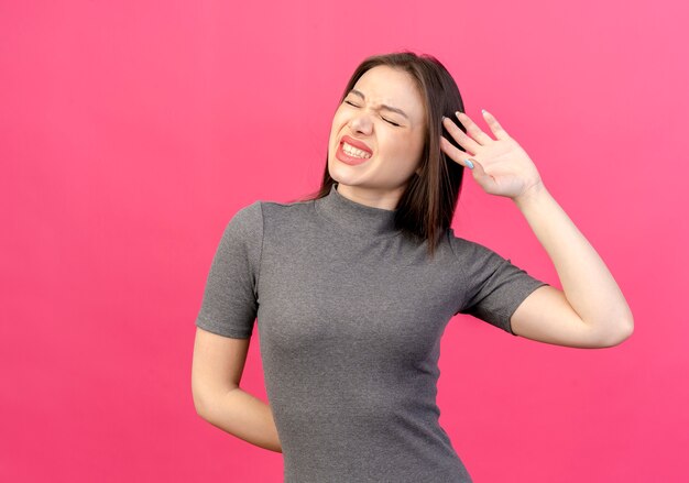 Mujer bonita joven dolorida manteniendo la mano detrás de la espalda y tocando la cabeza con la otra mano con los ojos cerrados aislado sobre fondo rosa con espacio de copia