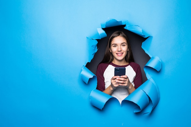 Mujer bonita joven escribiendo y usa el teléfono mientras mira a través del agujero azul en la pared de papel.