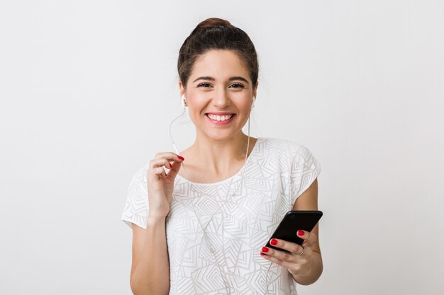 Mujer bonita joven con estilo sonriendo en camiseta, escuchando música en auriculares, sosteniendo el teléfono inteligente, usando el dispositivo, gesto aislado, feliz y positivo