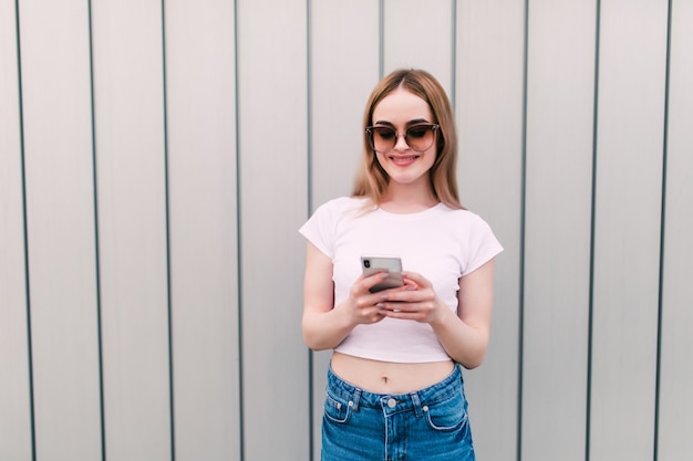 La mujer bonita joven en gafas de sol utiliza el teléfono móvil que se opone a la pared rayada al aire libre