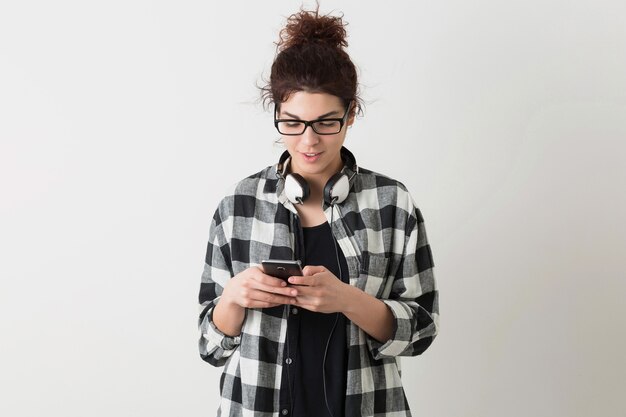 Mujer bonita joven con gafas, sosteniendo un teléfono inteligente, usando un dispositivo digital, sonriendo, feliz, auriculares, aislado sobre fondo blanco, camisa a cuadros, estilo hipster, estudiante, escribiendo mensaje
