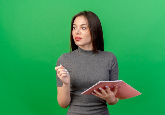 Mujer bonita joven mirando al lado sosteniendo el bloc de notas y la pluma