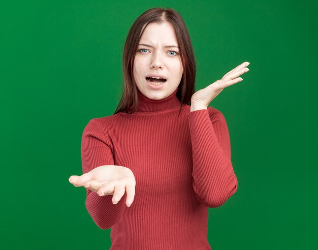 Mujer bonita joven molesta mirando al frente mostrando las manos vacías aisladas en la pared verde