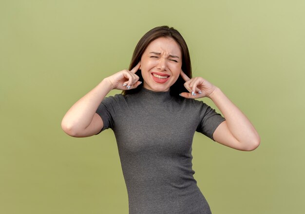 Mujer bonita joven molesta poniendo los dedos en los oídos con los ojos cerrados aislado sobre fondo verde oliva