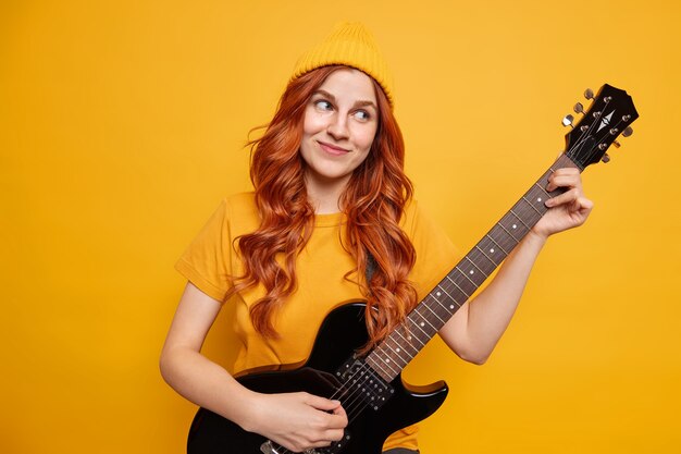 Mujer bonita joven con el pelo rojo disfruta de tocar la guitarra acústica tiene expresión complacida de ensueño