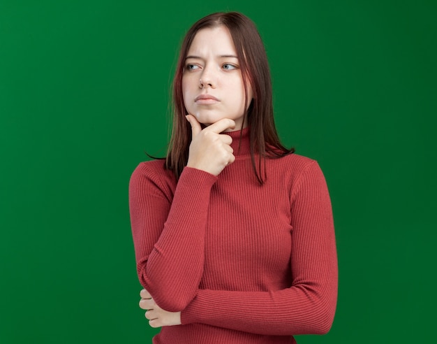 Mujer bonita joven pensativa manteniendo la mano en la barbilla mirando al lado aislado en la pared verde con espacio de copia