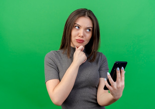Mujer bonita joven pensativa que sostiene el teléfono móvil que toca la barbilla y que mira el lado aislado en fondo verde con el espacio de la copia