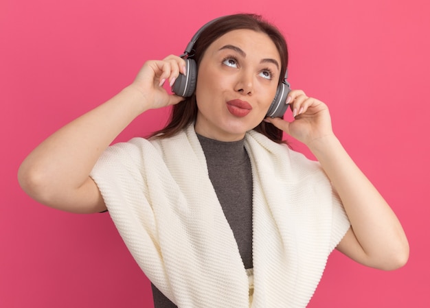 Mujer bonita joven pensativa que usa y que agarra los auriculares que miran hacia arriba aislados en la pared rosada