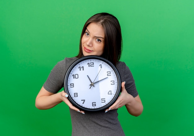 Mujer bonita joven pensativa sosteniendo el reloj mirando al lado aislado sobre fondo verde con espacio de copia