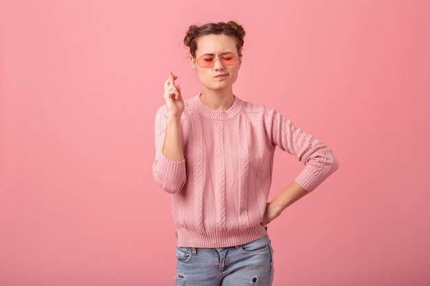 Mujer bonita joven pidiendo un deseo cruzando los dedos en suéter rosa y gafas de sol aisladas sobre fondo rosa studio