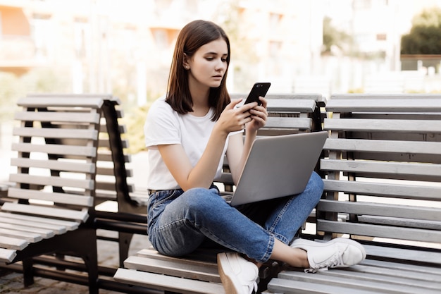 Mujer bonita joven que se sienta en banco en parque verde el día de verano y que lee el mensaje de texto en el teléfono celular mientras que usa la computadora portátil de plata.