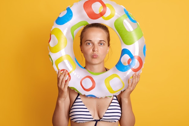 Foto gratuita la mujer bonita joven que se siente cansada y frustrada, que parece frustrada con el problema, que presenta con los labios fruncidos, que sostiene el anillo inflable delante de la cara, se coloca contra la pared amarilla.