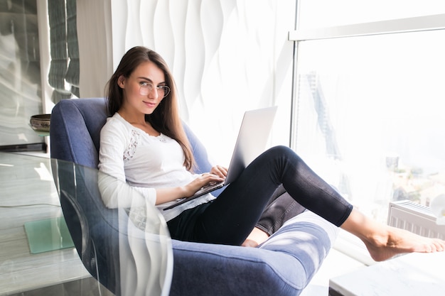 Mujer bonita joven que trabaja en la computadora portátil mientras está sentado en la silla en casa nueva ventana de luz