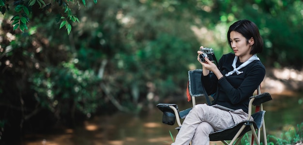 Mujer bonita joven que usa una cámara digital para tomar una foto de la hermosa naturaleza mientras acampa en el bosque con espacio de copia de felicidad