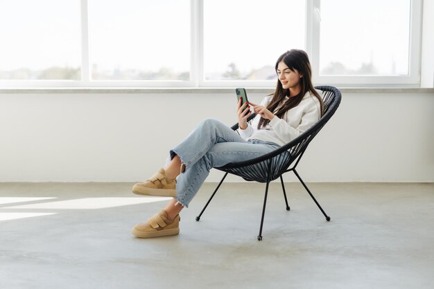 Mujer bonita joven que usa el teléfono móvil mientras está sentada en un sillón acogedor