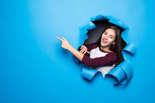 Foto gratuita la mujer bonita joven señaló el lado mientras que miraba a través del agujero azul en la pared de papel.