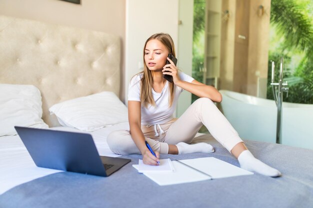 Mujer bonita joven sentada en la cama con su computadora portátil y hablando por teléfono con el bloc de notas alrededor