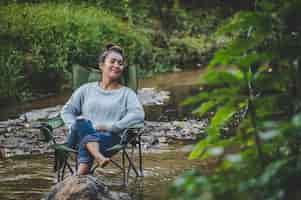 Foto gratuita mujer bonita joven sentada en una silla de camping en el arroyo y cierra los ojos para relajarse, sonríe en el bosque natural mientras acampa con espacio de copia de felicidad