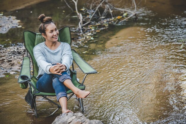 Mujer bonita joven sentada en una silla de camping en el arroyo para relajarse, sonríe en el bosque natural mientras acampa con espacio de copia de felicidad