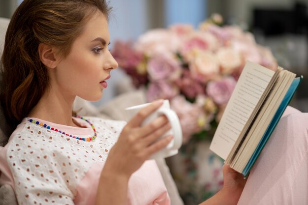 Mujer bonita joven sentada en el sofá tomando café y leyendo un libro disfruta del descanso