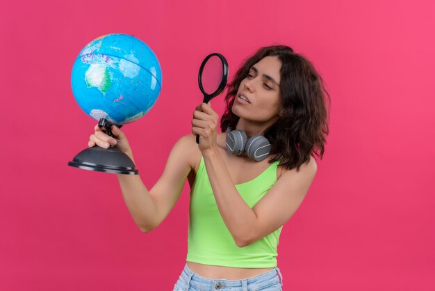 Una mujer bonita joven seria con el pelo corto en la parte superior verde de la cosecha en auriculares mirando el mundo constantemente con lupa