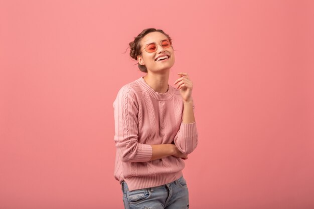 Mujer bonita joven con sincera sonrisa sincera en suéter rosa y gafas de sol aisladas sobre fondo rosa studio