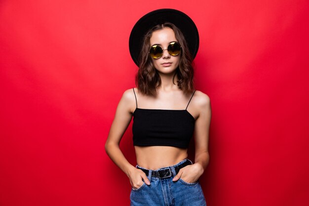 Mujer bonita joven en sombrero de ala ancha y gafas de sol que se encuentran aisladas en la pared roja