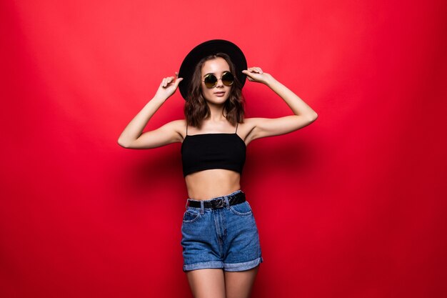 Mujer bonita joven en sombrero de ala ancha y gafas de sol que se encuentran aisladas en la pared roja