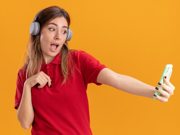 Mujer bonita joven sorprendida en auriculares sostiene y mira el teléfono aislado en la pared naranja