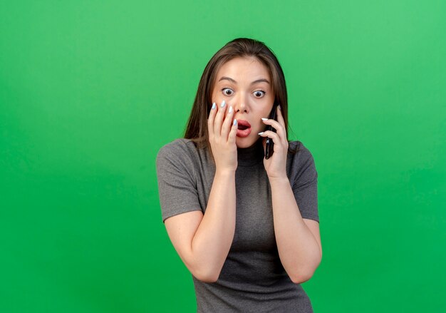 Mujer bonita joven sorprendida mirando directamente hablando por teléfono tocando la cara aislada sobre fondo verde con espacio de copia